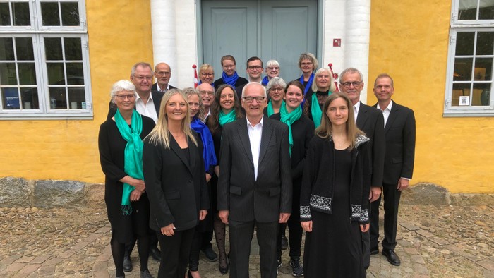 En gruppe af formelle personer står sammen foran et gult og hvitt hus.