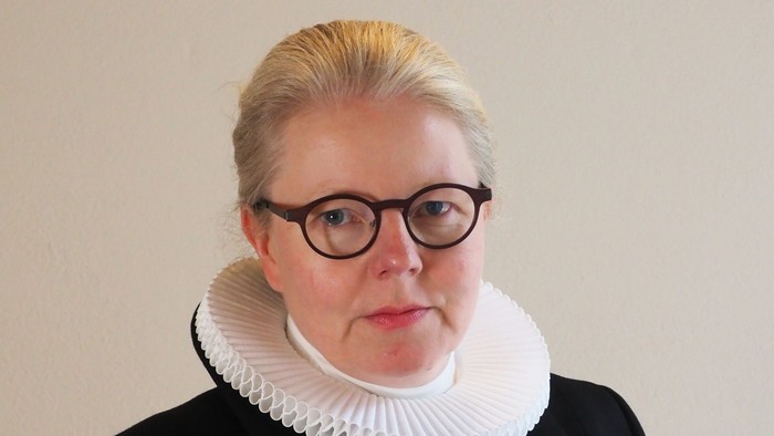 En kvinde med blond hår og briller bærer en sort jakke med hvid krage.