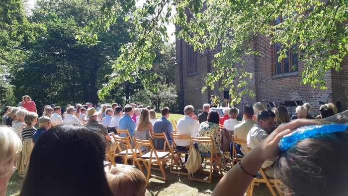 Eine große Menschenmenge sitzt auf Holzbänken vor einem Backsteingebäude im Freien.