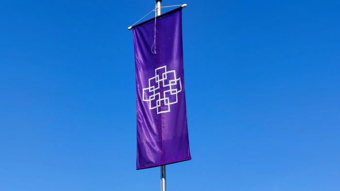 Ein hoch montierter lila Fahne mit weißem Kreuz vor grünen Bäumen und blauem Himmel