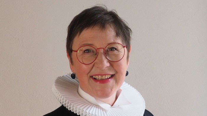 En kvinde med kort hår og røde briller, smiler og har et hvidt rødt krage under en sort jakke