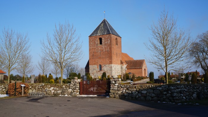 En rød murstenskirke med tårn bagved en murstengevel og træer