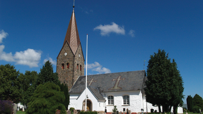 En lille kirke med tårn og gravhøje på en grøn plads under et blødt blå himmel.