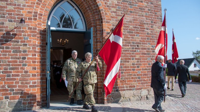 En gruppe uniformerede mænd går ud af et murstensbygning med danske flag.
