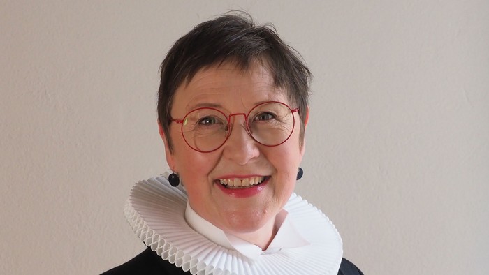 En kvinde med kort hår og røde briller, smiler og bærer en sort jakke med hvid krage.