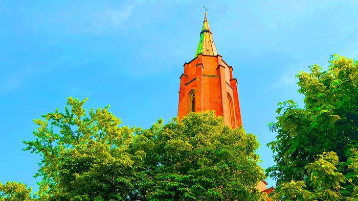 Ein roter Kirchturm ragt hinter grünen Bäumen in den blauen Himmel