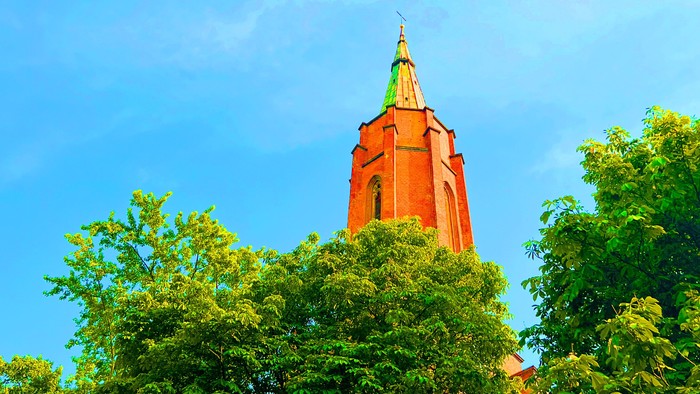 Ein roter Kirchturm ragt hinter grünen Bäumen in den blauen Himmel