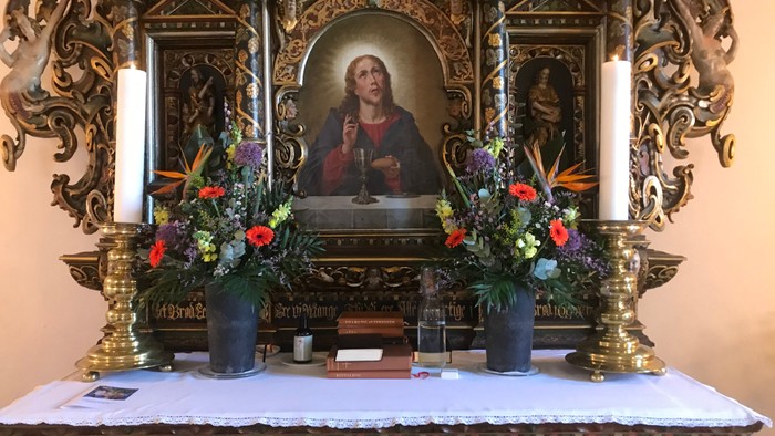 En præst preker i en præktig, udsmykket kirkealter med blomsterarrangeringer og lysstake.