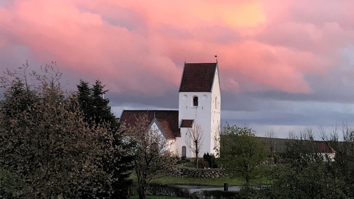 En hvid kirke med tårn og spiret mod en rosa skyer og solnedgang