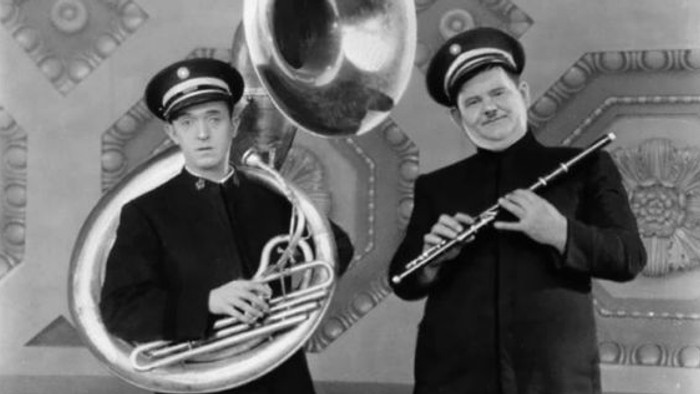 Zwei Männer in historischen Uniformen spielen Tuba und Flöte vor einem kunstvoll verzierten Hintergrund.