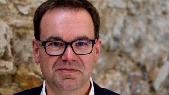 Mann mit Brille und Anzug vor steinerner Wand.