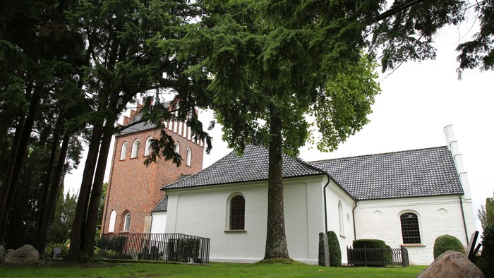 Et hvidt kirkebygning med sort tag og en højtårnet, omgivet af træer og græs.