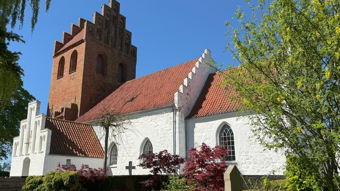 En hvid kirke med røde tag og tårn, omgivet af grønne buske og træer