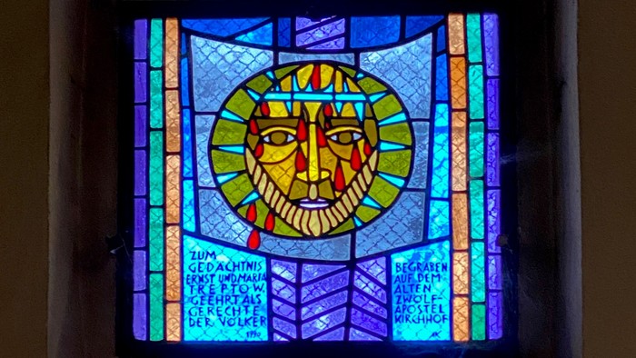 Buntglasfenster mit einem ruhigen Gesicht und einem sonnenähnlichen Symbol, umgeben von lebendigen Farben und architektonischen Linien.