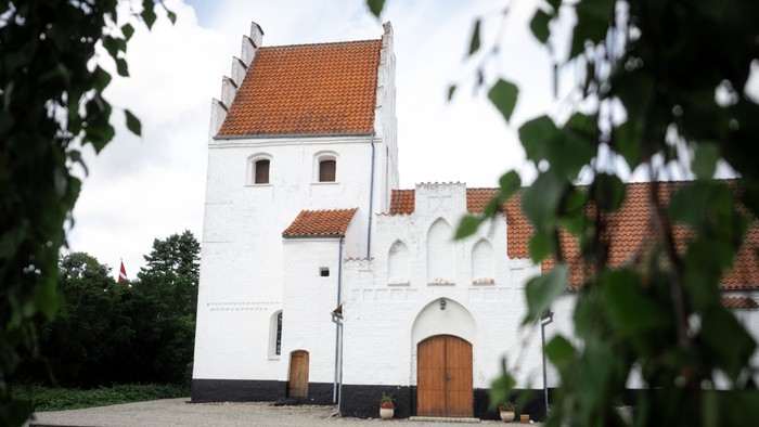 Et hvidt historisk tårn med røde tagfliser og trædør, omgivet af grønne buske og en benk.