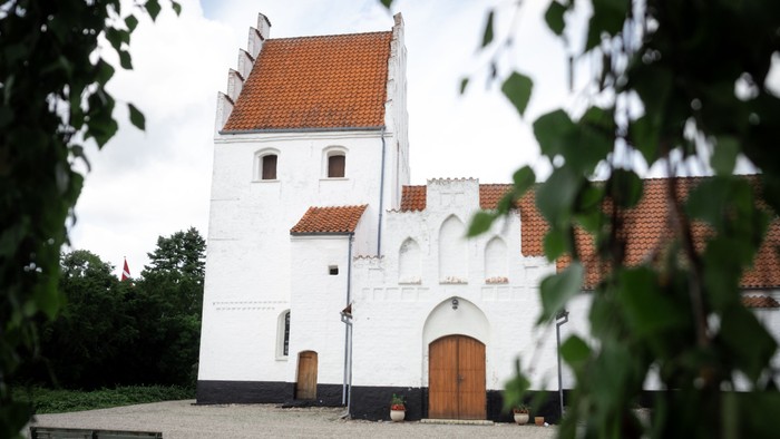 Et hvidt historisk tårn med røde tagfliser og trædør, omgivet af grønne buske og en benk.