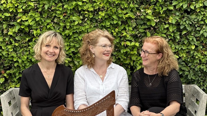 Tre kvinder sidder på en bænk, hvor den midterste spiller en harpe