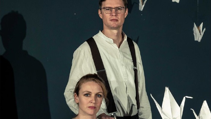 Ein Mann und eine Frau posieren vor einer blauen Wand mit Origami-Kranichen.