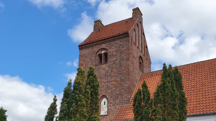 En rød murstenskirke med spiret og klokkehuset, omgivet af grønne træer og græs.