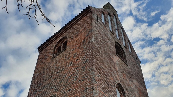 En høj murstensklokketårn med små vinduer og spids tak mod en blå Himmel med hvidt og grå skyer.