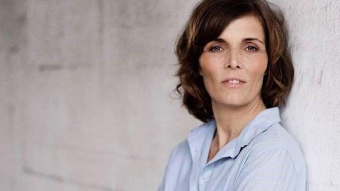 En kvinde med kort, brunt hår og blå bluse står foran en betonmur.