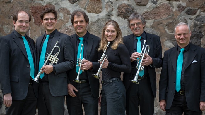 Eine Gruppe von sechs Musikern posiert mit Blechblasinstrumenten vor einer Steinmauer.