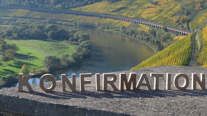 Ein Panorama-Blick auf eine Weinberglandschaft mit einem Fluss und einer Brücke, darunter ein Schild mit der Aufschrift "KONFIRMATION".
