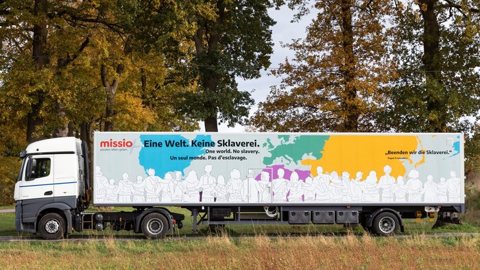 Ein weißer LKW mit buntem Design steht auf einem Feld, umgeben von Bäumen.