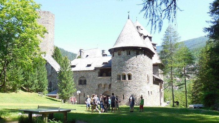 Ein Steinhaus mit Turm, umgeben von Bäumen und Menschen auf der Wiese