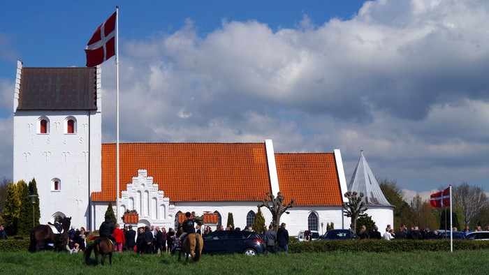 En gruppe heste og ryttere foran et hvidt og orangenet kirkebygning med dansk flag.