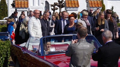 En gruppe mennesker omgiver en klassisk bil på gaden foran et hus med rødt tag.