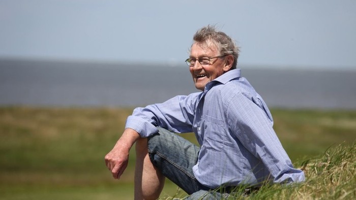 En glad mand sitter på en hæk i naturen og kigger ud over en strand.