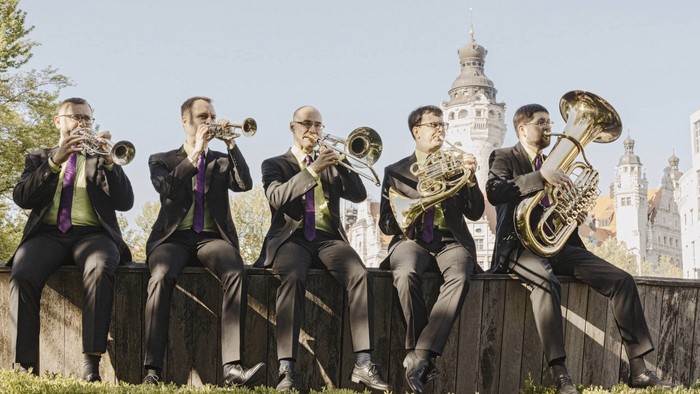 Eine Gruppe von Musikern sitzt auf einer Mauer und spielt Blechblasinstrumente vor einem Schloss.