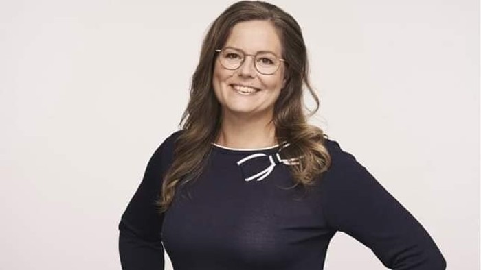 En kvinde med briller og langt mørkt hår, smiler og har armene krydset.