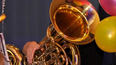 Eine Person spielt ein großes goldenes Tuba-Instrument, umgeben von bunten Ballons.