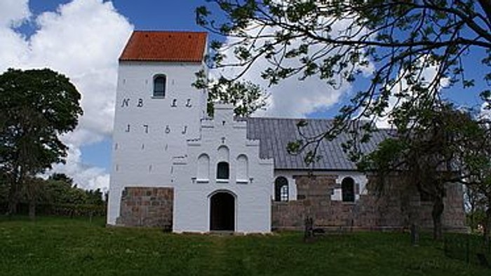 Et hvidt historisk kirkebygning med rødt tag og en mur til, beliggende i grønne omgivelser