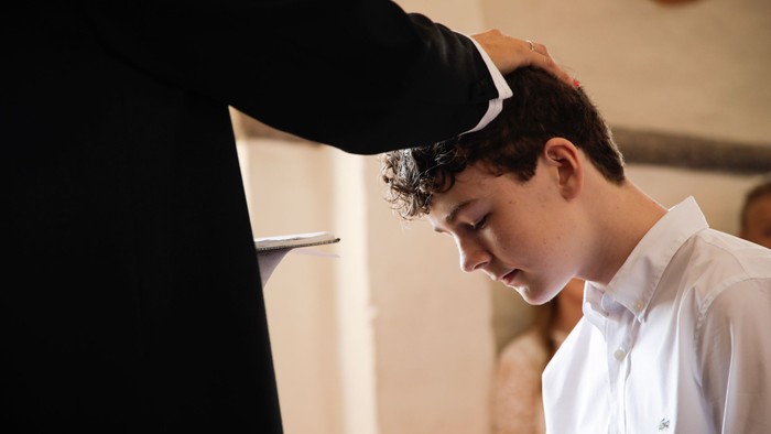 En person i formelt tøj velsigner en ung mand under en religiøs ceremoni.