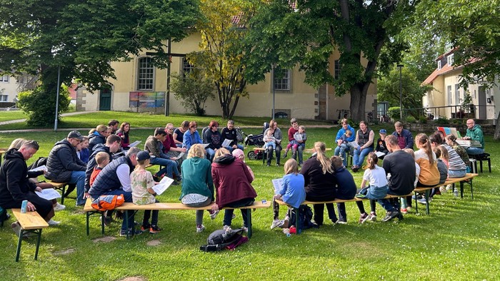 Eine Gruppe von Menschen sitzt im Freien auf Bänken und Stühlen auf einem grasbewachsenen Bereich, umgeben von Bäumen und Gebäuden.