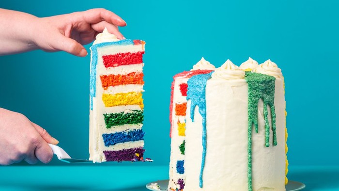 Eine Hand hält ein Stück Regenbogenkuchen neben einem ganzen Kuchen mit bunten Streifen.