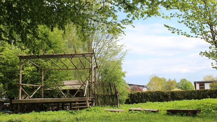 Ein hölzerner Pavillon in einem grünen Garten mit Bäumen und Büschen.