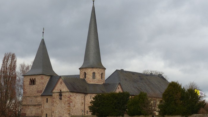Historische Steinkirche mit Zwillingstürmen vor bewölktem Himmel und umgeben von Grün.