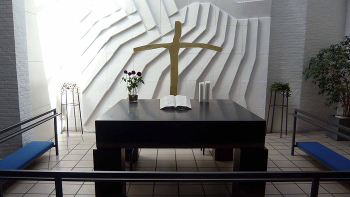 En slem med blomster og kande foran et stort kors på en muret vegg