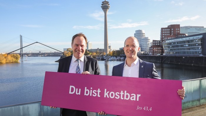 Zwei Männer halten ein pinkfarbenes Schild mit dem deutschen Text *„Du bist kostbar“* (Du bist wertvoll) vor einer Flussufer-Stadtkulisse.