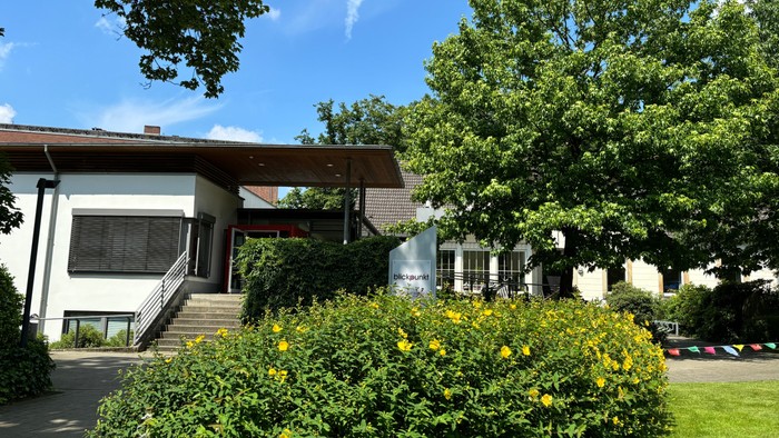Hier sieht man das Gemeindehaus blick.punkt, ein weißes, flaches Gebäude mit großer Treppe und einer roten Tür.