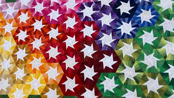 Eine Patchworkarbeit in Regenbogenfarben, viele Sechsecke von gelb über orange, rot, blau und grün, immer mit einem weißen Rad in der Mitte.