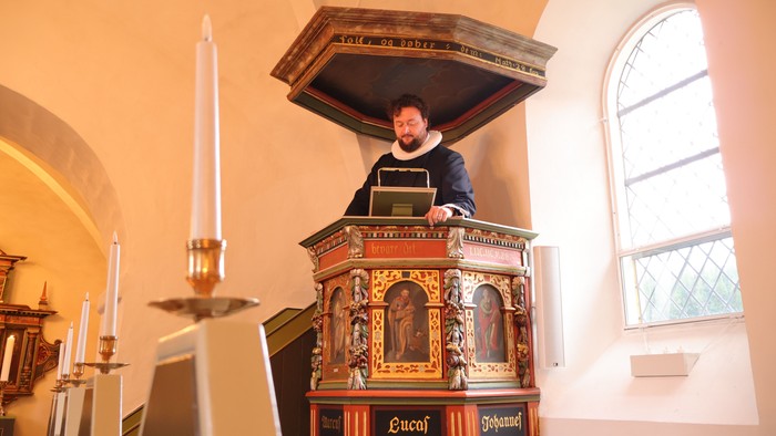 En person står ved en prægtig prædikestol i en kirke og læser op fra en bog under en gudstjeneste.