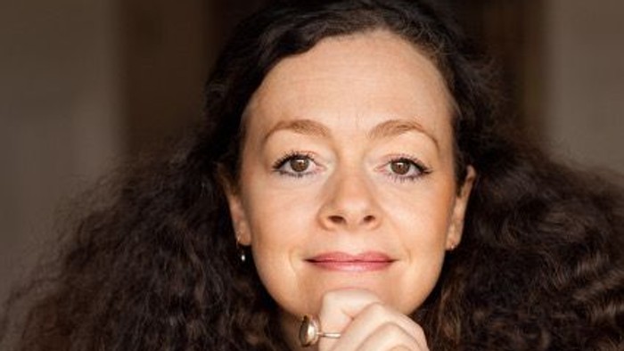 Eine Person mit lockigem Haar posiert nachdenklich, das Kinn entspannt auf die Hand gestützt.