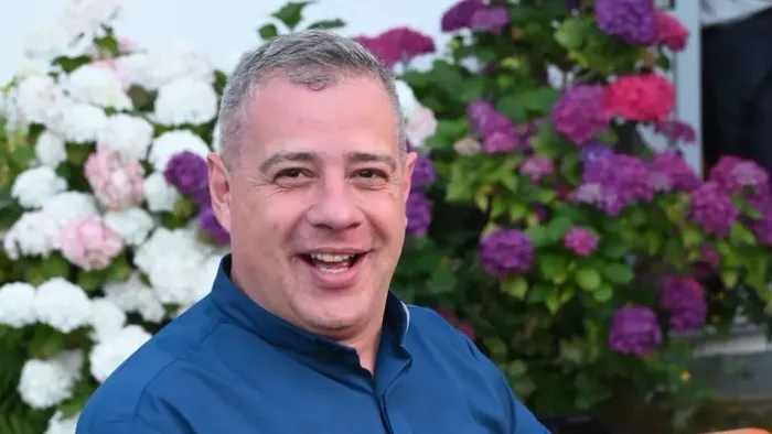 Ein lächelnder Mann in einem blauen Hemd posiert neben üppigen blühenden Sträuchern im Freien.