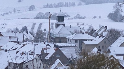 Schneebedecktes Dorf mit einem markanten Gebäude in der Mitte, umgeben von Feldern und Bäumen.