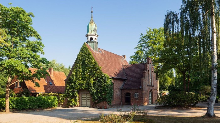 Kleine Kirche mit grünem Turm und rotem Dach, umgeben von Bäumen und Büschen